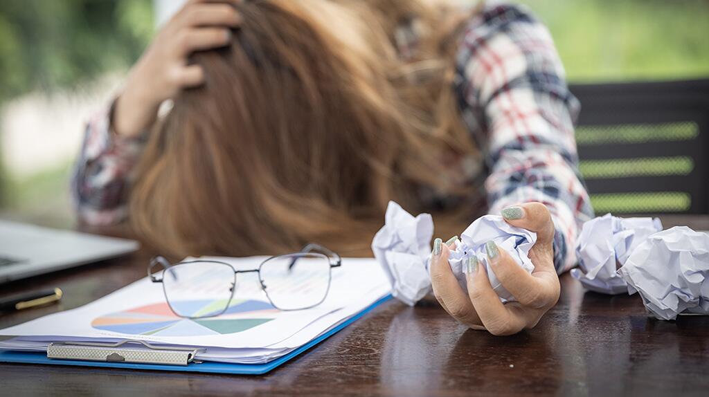 Vrouw met hoofd in handen naast bril en verfrommeld papier op tafel.