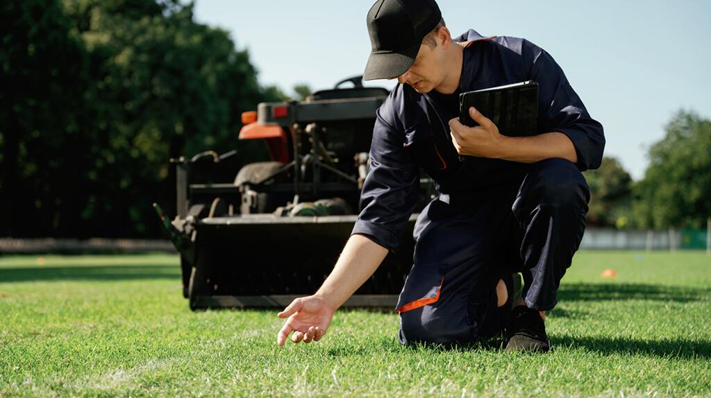 Man knielt op gras naast grasmaaier, inspecteert het veld.