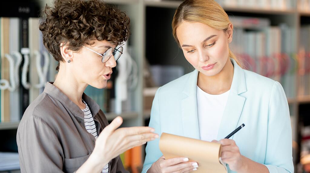 Twee vrouwen overleggen in een kantoor, de een maakt notities.