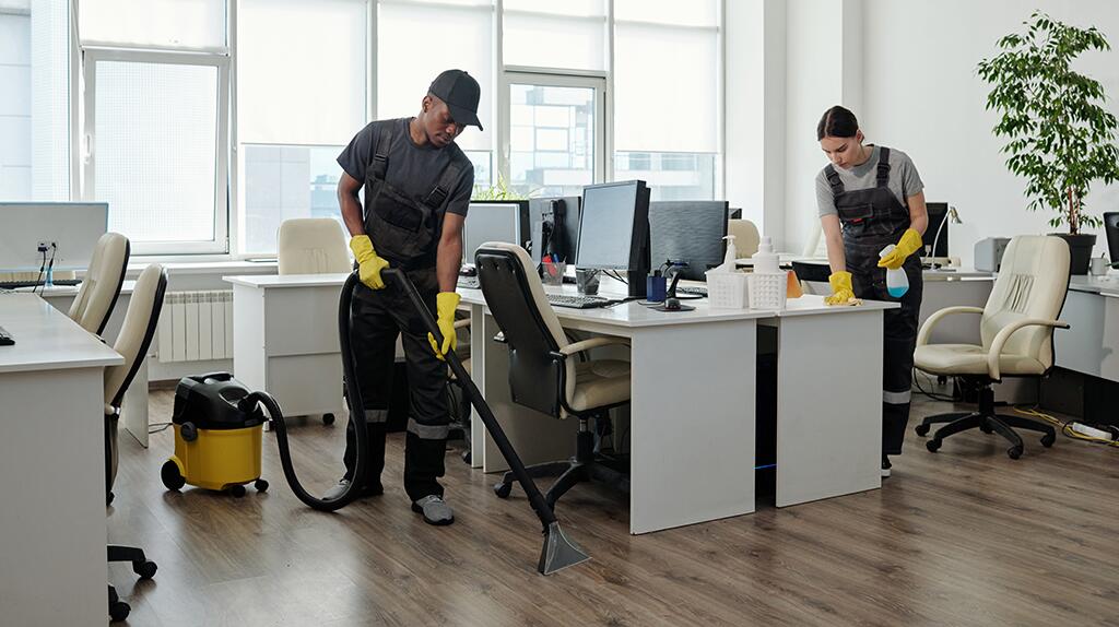 Schoonmakers stofzuigen en poetsen een modern kantoor met bureaus en computers.
