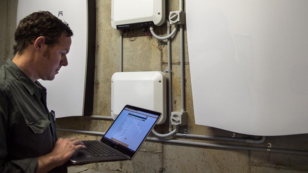 Man werkt op laptop naast opgehangen apparaten aan muur.