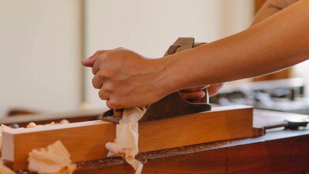 Houten plank wordt geschaafd door handen met een schaaf, houtkrullen zichtbaar.