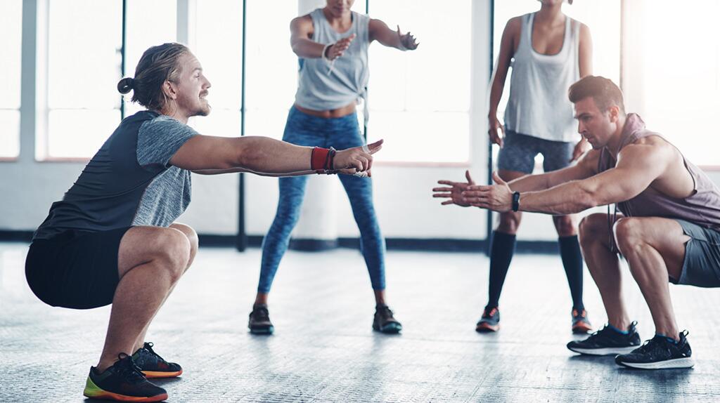 Twee mannen doen squats tegenover elkaar in een sportschool; twee vrouwen op de achtergrond.