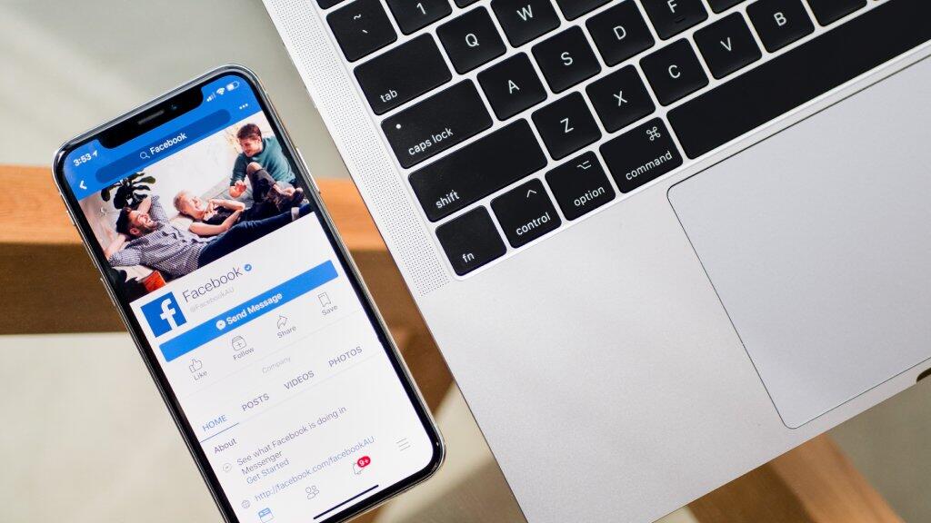 Smartphone met Facebook-app naast laptop op houten tafel.