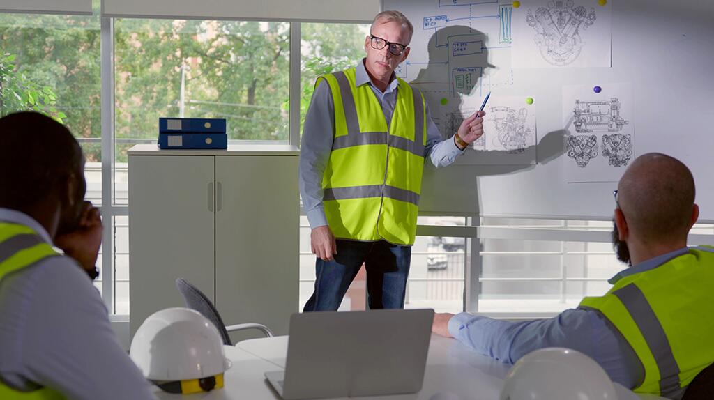 Man in veiligheidshes presenteert aan groep, wijzend naar bouwtekeningen op muur.