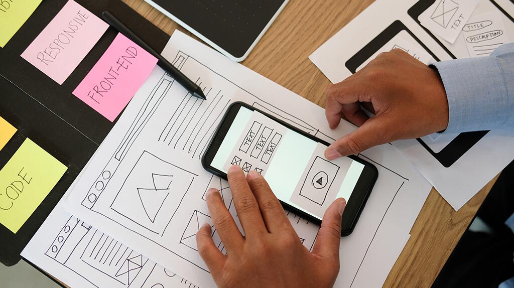 Handen werken op een smartphone met ontwerpuitdraaien en kleurige notities op tafel.