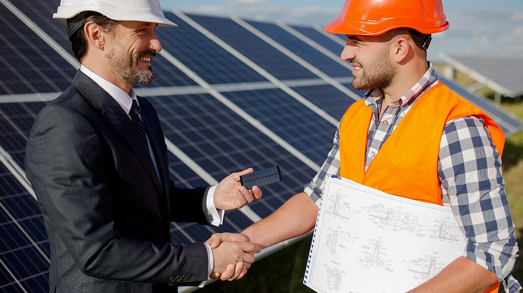 Twee mannen schudden handen bij zonnepanelen, beiden dragen helmen.