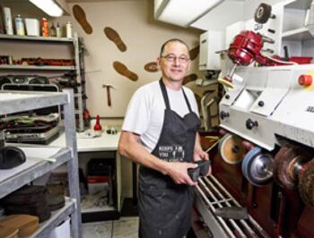 Schoenmaker aan het werk in een werkplaats, lachend naar de camera.