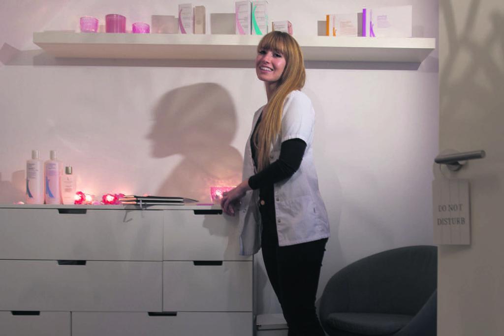 Vrouw in een witte jas in een verzorgingskamer, glimlachend naast een kast met producten.