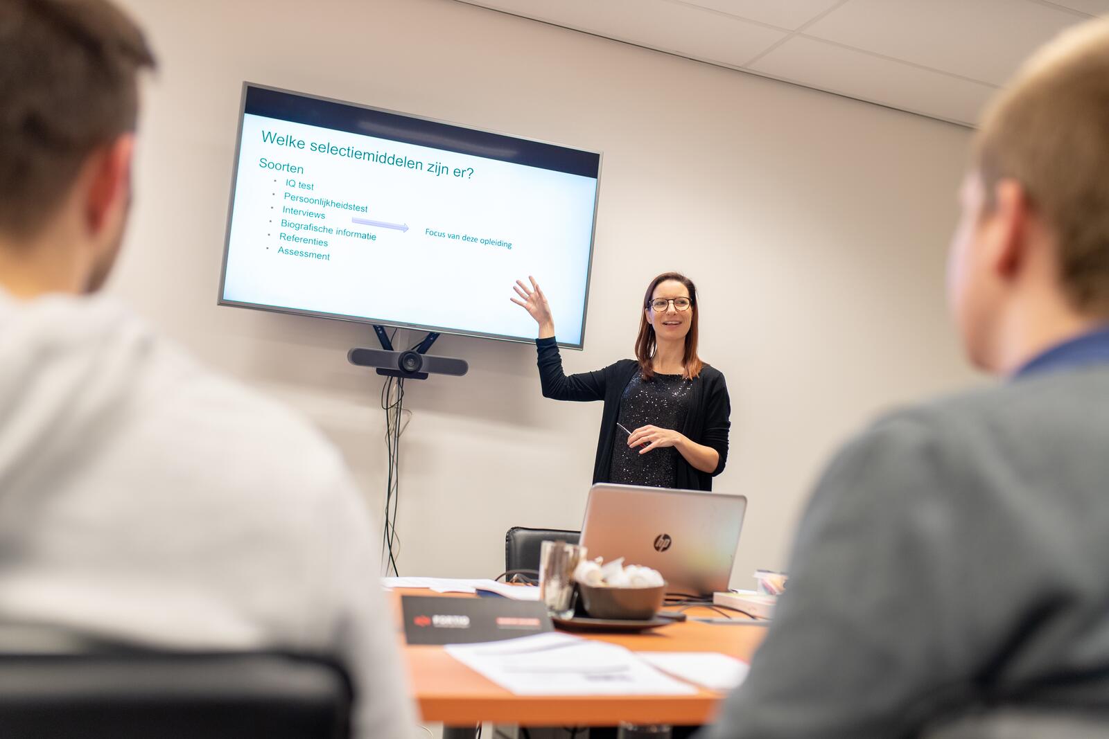 Vrouw geeft presentatie aan collega’s met een laptop en scherm in vergaderruimte.