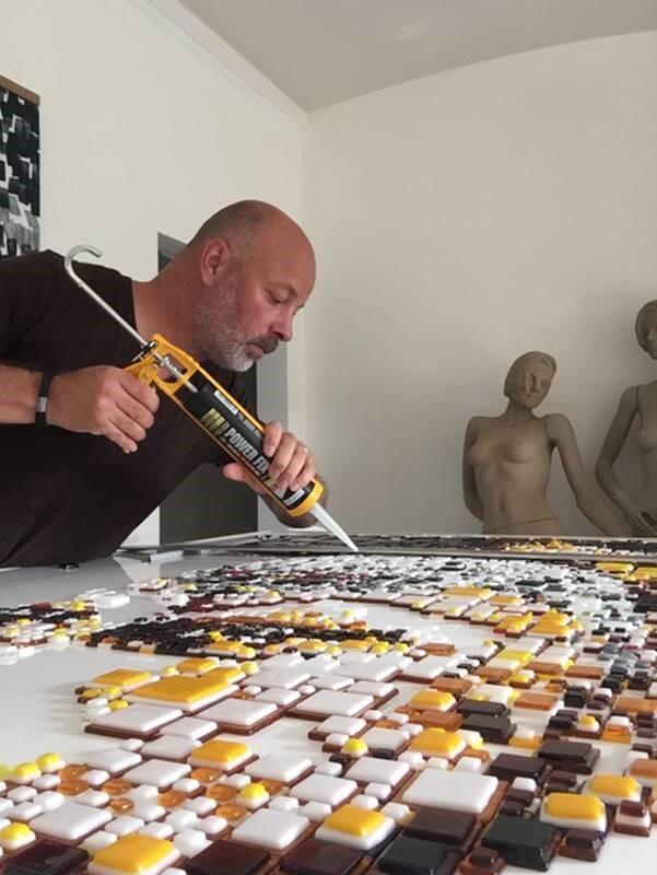 Man werkt met tegels en lijmpistool in lichte kamer met sculpturen.