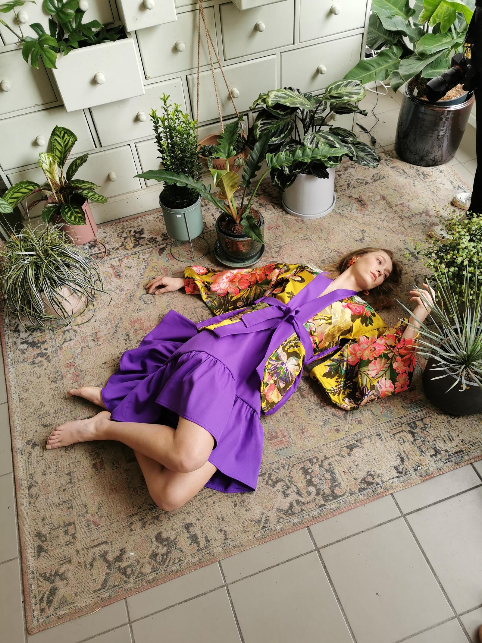 Vrouw in paarse jurk ligt ontspannen op een tapijt omringd door groene planten.