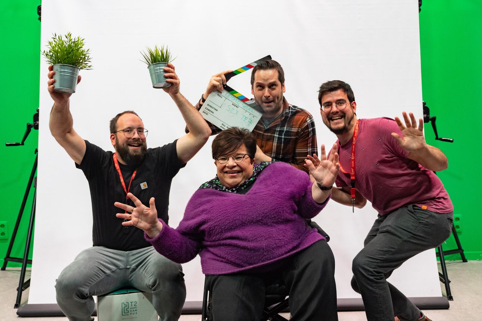 Vier lachende mensen poseren met planten en een klapbord voor een groene en witte achtergrond.