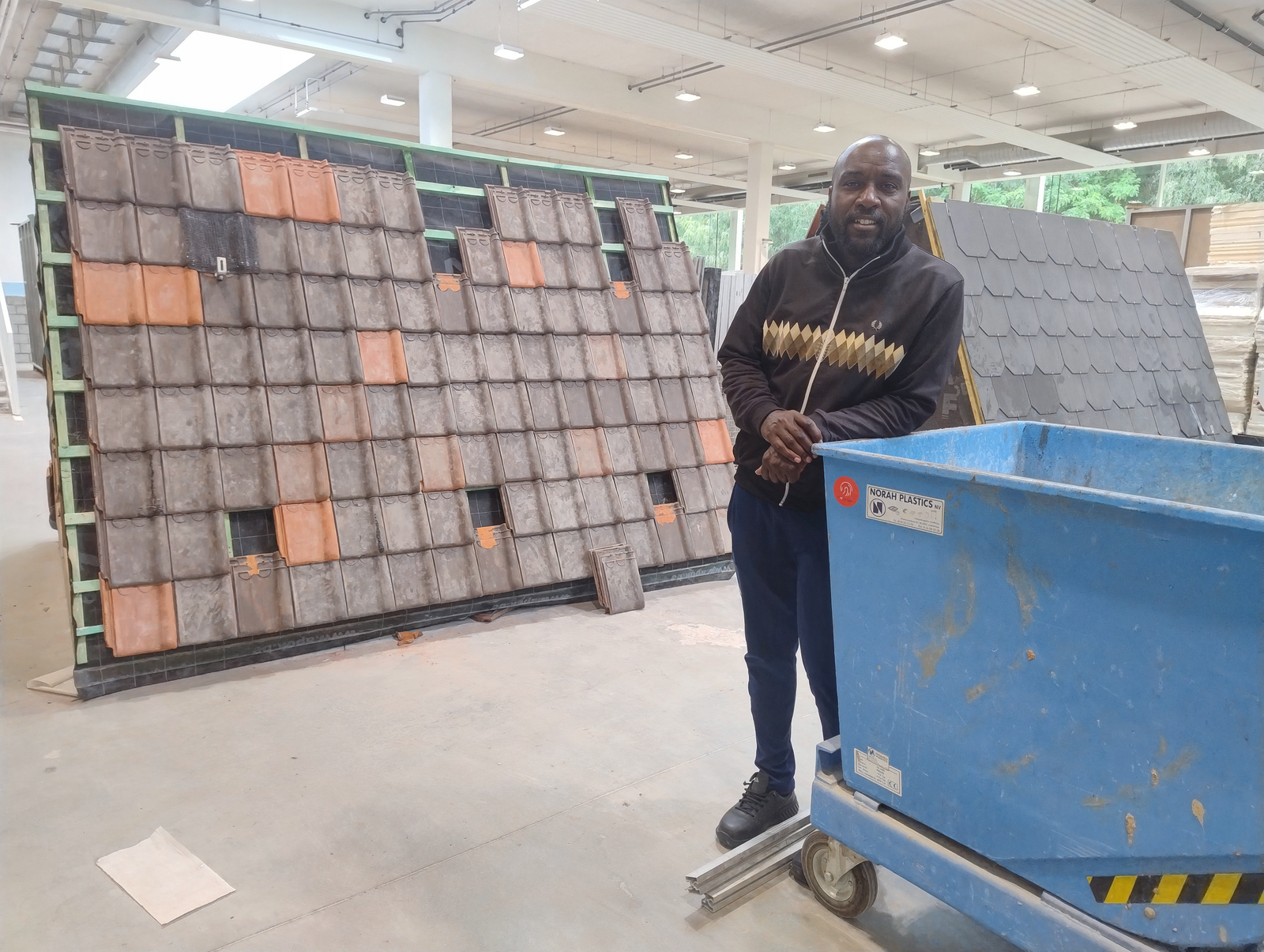 Man poseert bij dakpannenmuur en blauwe container in werkplaats.