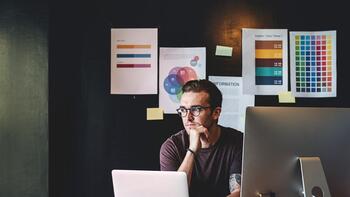 Man werkt aan computer, nadenkend bij bureau, met grafieken op muur.