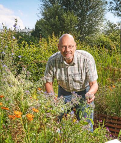 Man in tuin met groene planten en oranje bloemen, lachend naar de camera.