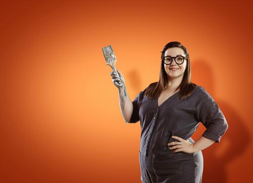 Vrouw met bril, grijs pak en prothesehand, lachend voor oranje achtergrond.