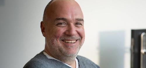 Glimlachende man met kaal hoofd en grijze trui, lichte achtergrond.