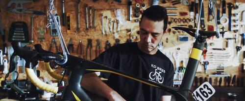 Man repareert fiets in werkplaats, omgeven door gereedschap.