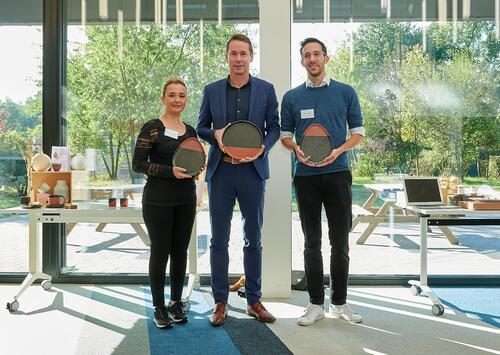 Drie mensen staan binnen met ronde awards in een zonnige ruimte.