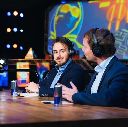 Twee mannen met headsets praten aan een tafel in een kleurrijke, verlichte studio.