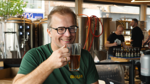 Man met bril glimlacht en houdt een glas donker bier vast in een bar.