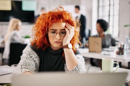 Vrouw met rood haar en bril kijkt vermoeid achter laptop in kantoor.