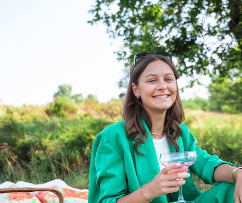 Vrouw in groene blazer lacht terwijl ze een drankje vasthoudt in een park.