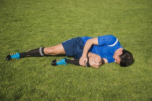 Voetballer in blauw liggend op gras, houdend zijn knie.