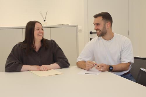 Twee mensen glimlachen en praten aan een tafel in een kantoorruimte.
