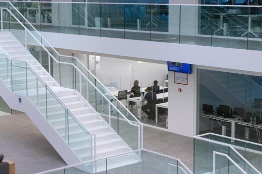 Binnenkant kantoor met glazen balustrades en grote trap, mensen werken aan bureaus.