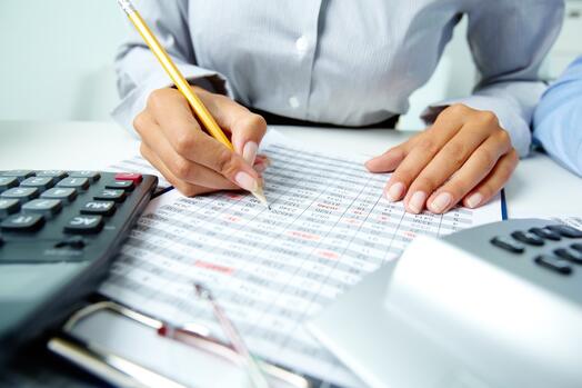 Persoon schrijft op een spreadsheet aan bureau met rekenmachine.