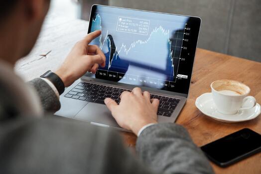 Man wijst naar aandelenverkoopgrafiek op laptop, met koffie en telefoon ernaast.