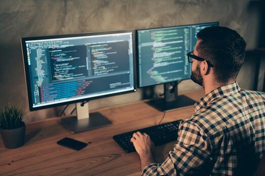 Man werkt aan computercode op twee schermen in een kantoor.