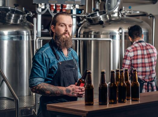 Man in brouwerij met fles, meerdere bierflessen op bar.