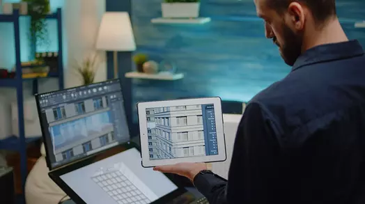 Man werkt op laptop en tablet in een modern kantoor, blauwe muren op de achtergrond.