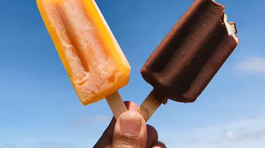 Twee ijslolly’s, eentje oranje en eentje bruin, tegen een blauwe hemel.
