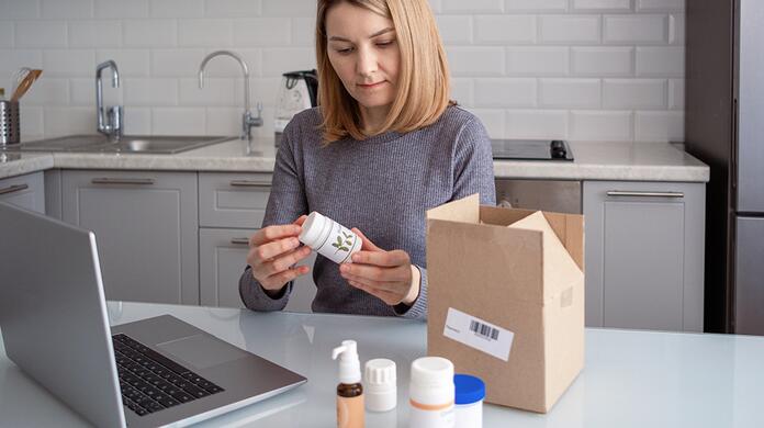 Vrouw onderzoekt medicijnpot in keuken, laptop en doos op tafel.