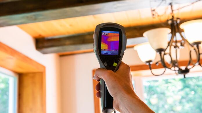 Hand houdt infraroodcamera in woonkamer; hout plafond, kroonluchter zichtbaar.