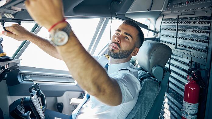 Piloot in de cockpit bedient instrumenten, zonlicht via ramen, brandblusser zichtbaar.