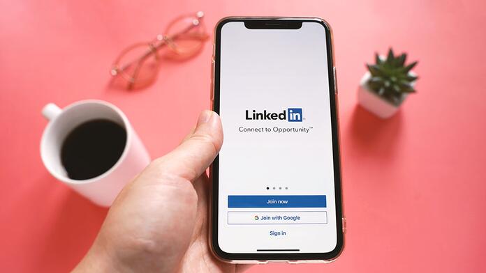 Hand houdt telefoon met LinkedIn-scherm vast, naast koffie, bril en plant.