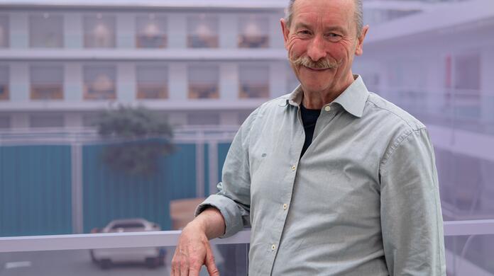 Man met grijze baard glimlacht voor een kantoorgebouw.