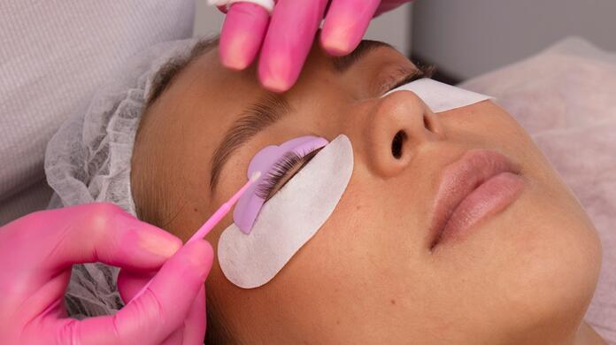 Wimperlifting met roze handschoenen en oogpads op een liggende vrouw.