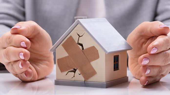 Twee handen beschermen een modelhuis met scheuren en pleister.