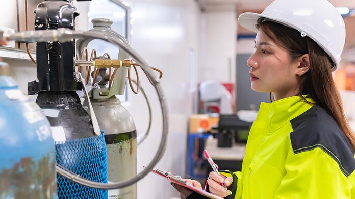 Vrouw in gele jas en helm inspecteert gasflessen in een industriële ruimte.