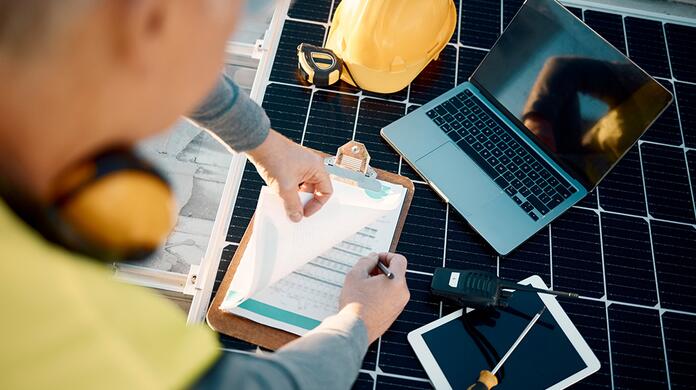 Man werkt met laptop en document op zonnepanelen, zonnebril en tablet naast hem.