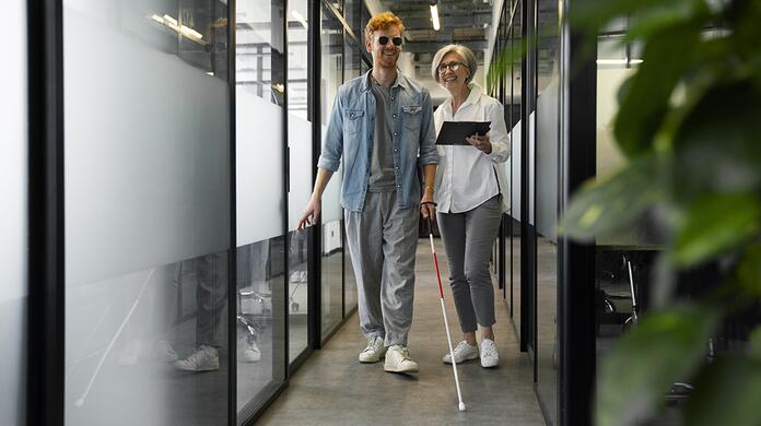 Twee mensen lopen met witte stok door een moderne gang.