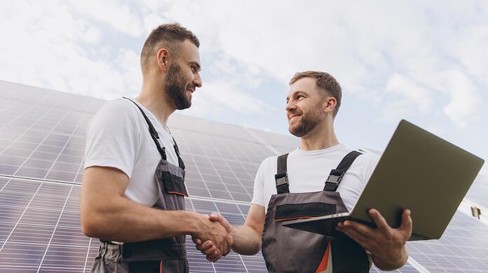 Twee mannen in overalls schudden handen bij zonnepanelen, één met laptop.