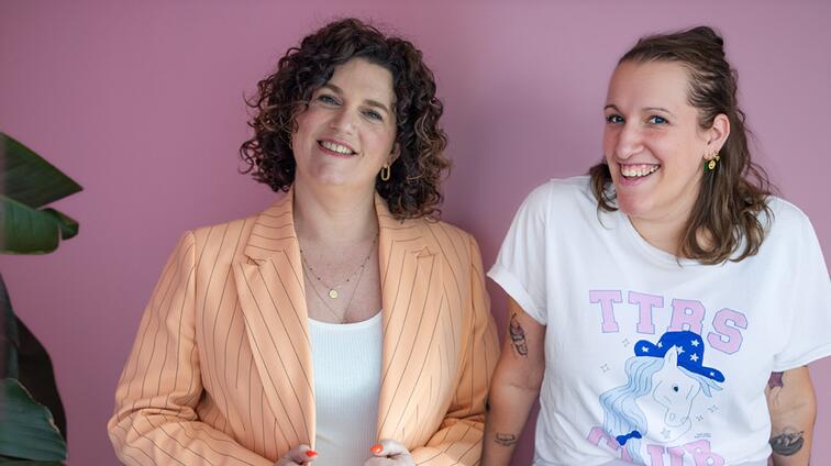 Twee lachende vrouwen tegen een roze achtergrond, één in oranje blazer, één in wit T-shirt.