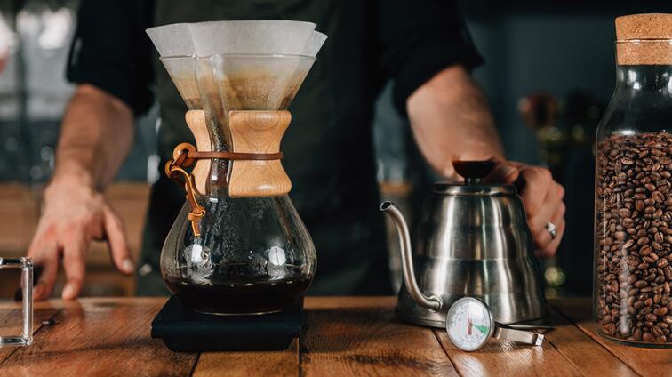 Hand bereidt koffie met Chemex en metalen waterkoker op houten tafel.
